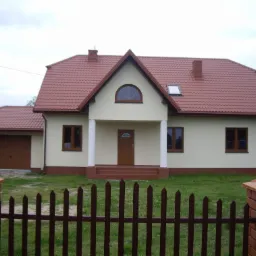 Nowo wybudowany dom jednorodzinny z garażem, wykończony w jasnych kolorach, z brązową stolarką okienną i dachem z czerwonej dachówki, widziany zza brązowego, sztachetowego ogrodzenia.